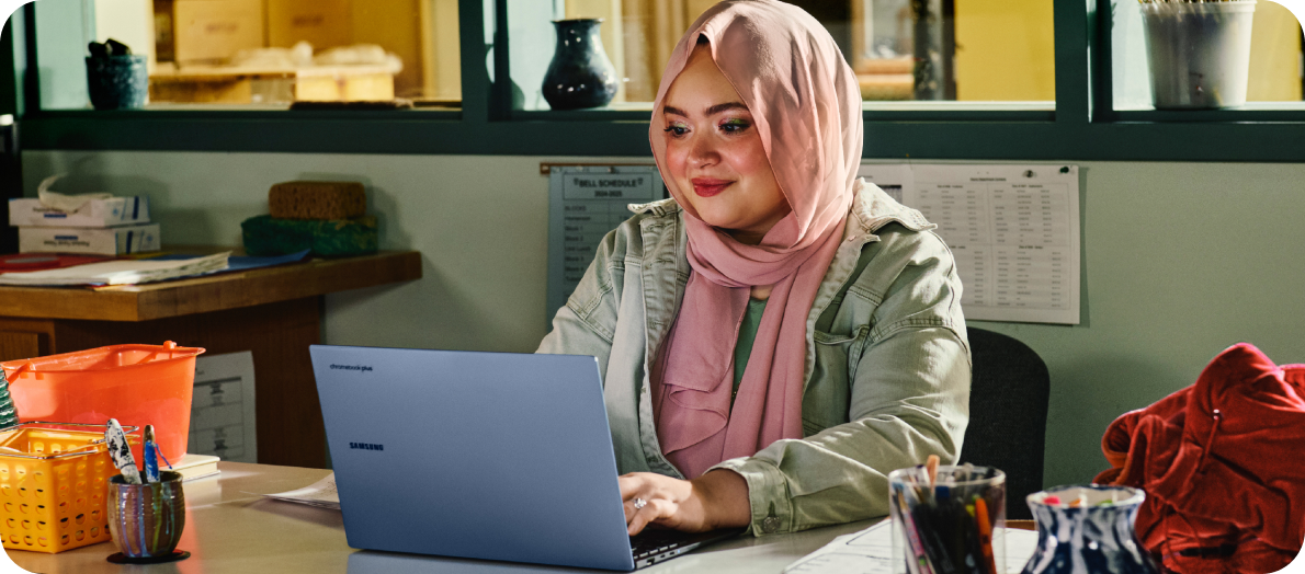 A smiling college student wearing a hijab sitting at a desk working on a Chromebook.